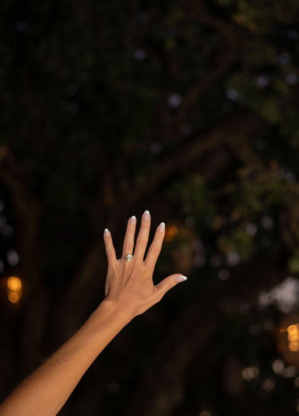 A hand showing an engagement ring in progressive stages of being raised against a dark outdoor evening background.