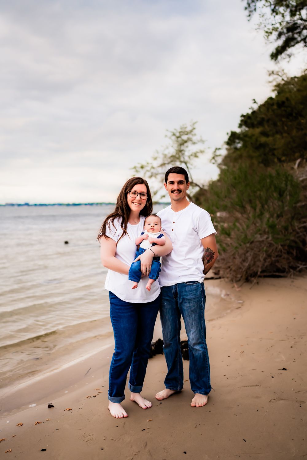 A family wearing matching white and denim outfits poses together on a beach at sunset with water in the background.