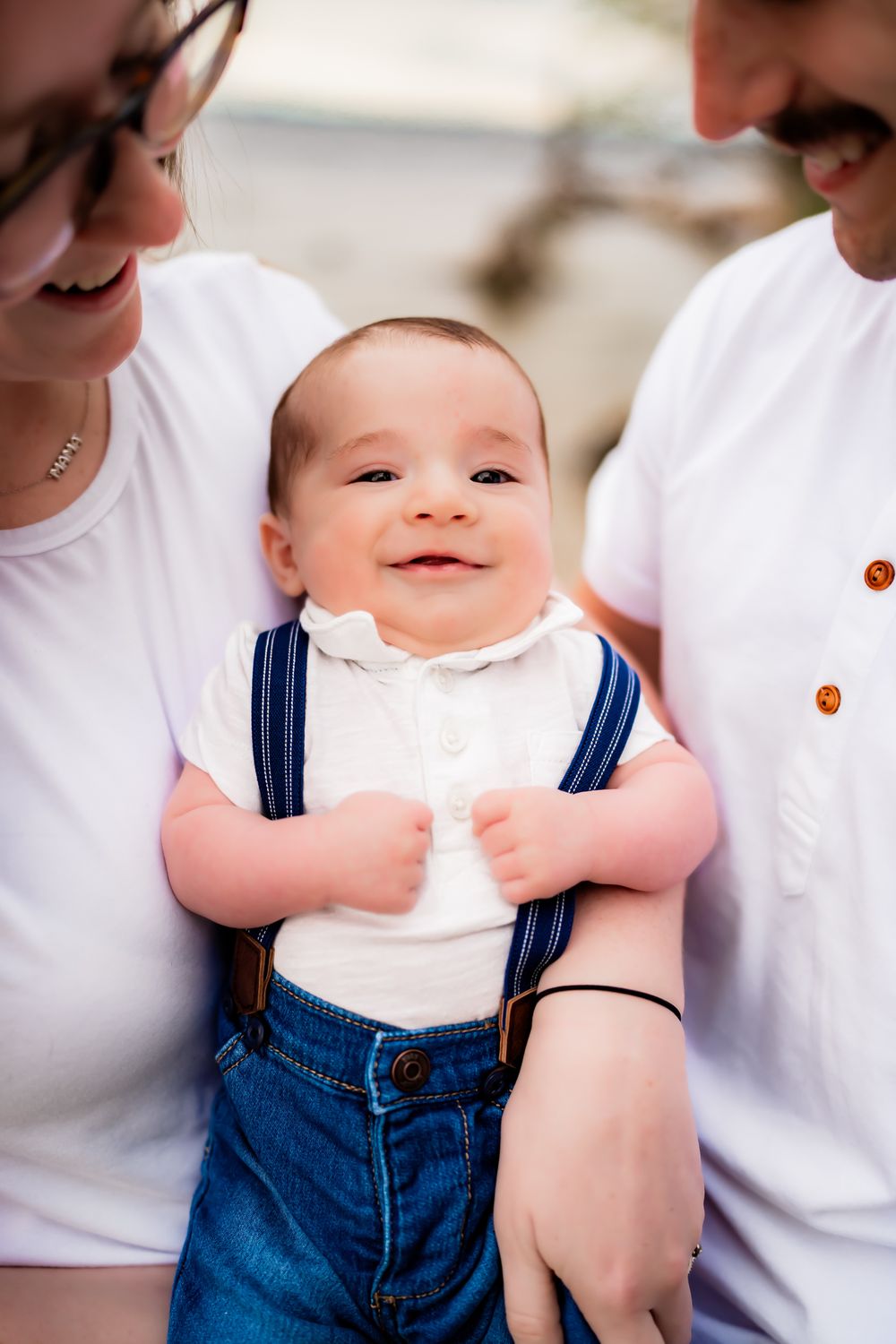 Baby wearing blue denim overalls and white shirt smiles joyfully against light background in close up photo series.
