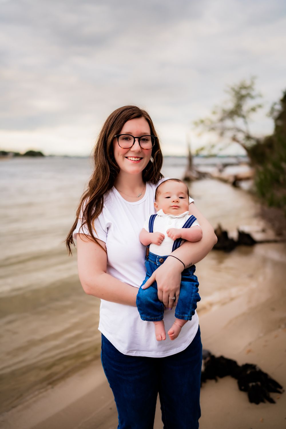 A parent and baby stand together on a beach at sunset with water and trees in the background.