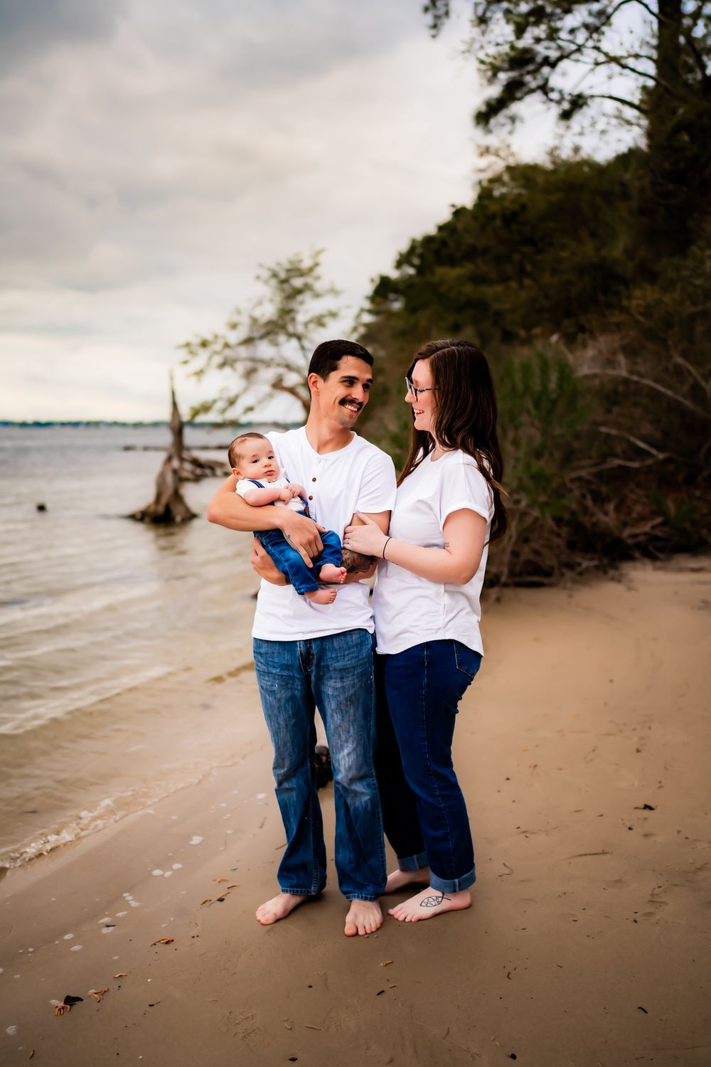 A family wearing matching white shirts and jeans poses together on a sandy beach at sunset.