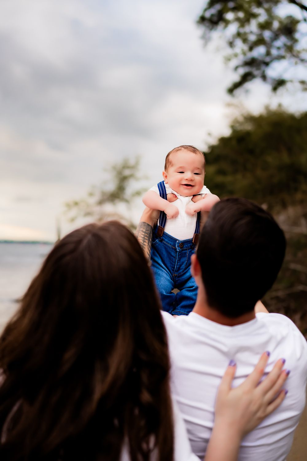 A happy baby smiles and laughs while being held up on a cloudy beach day.