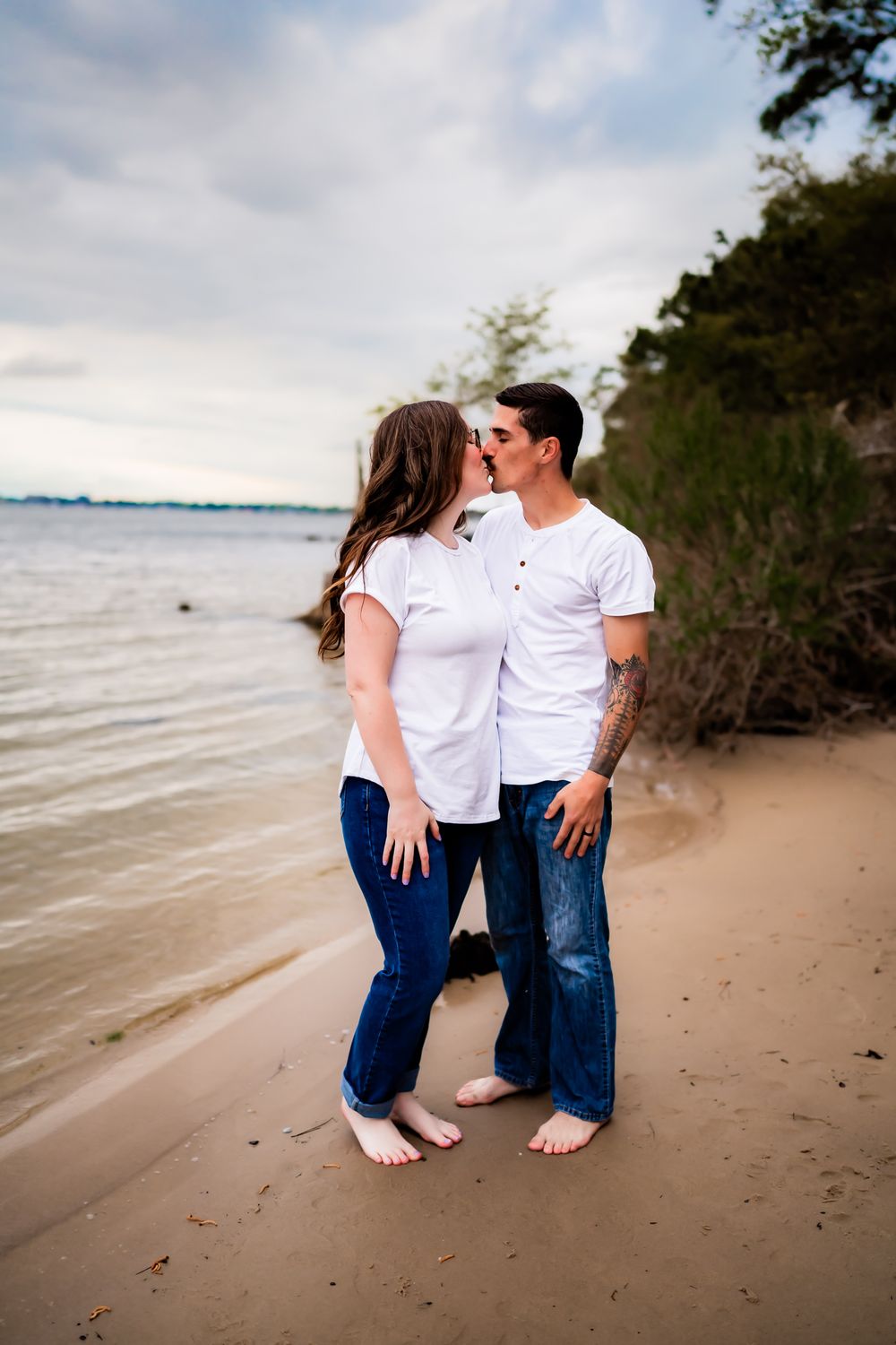 A couple in coordinating white shirts and jeans share an intimate moment on a beach shoreline.