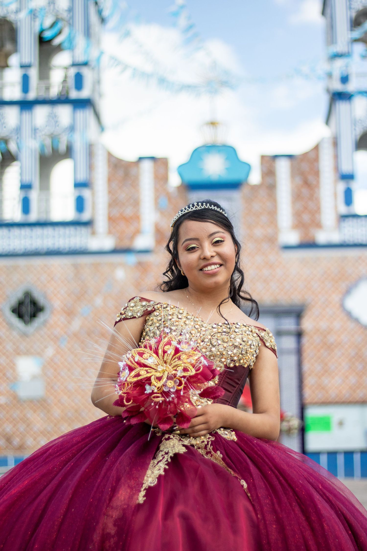 Young woman in burgundy and gold quinceanera gown holding bouquet.