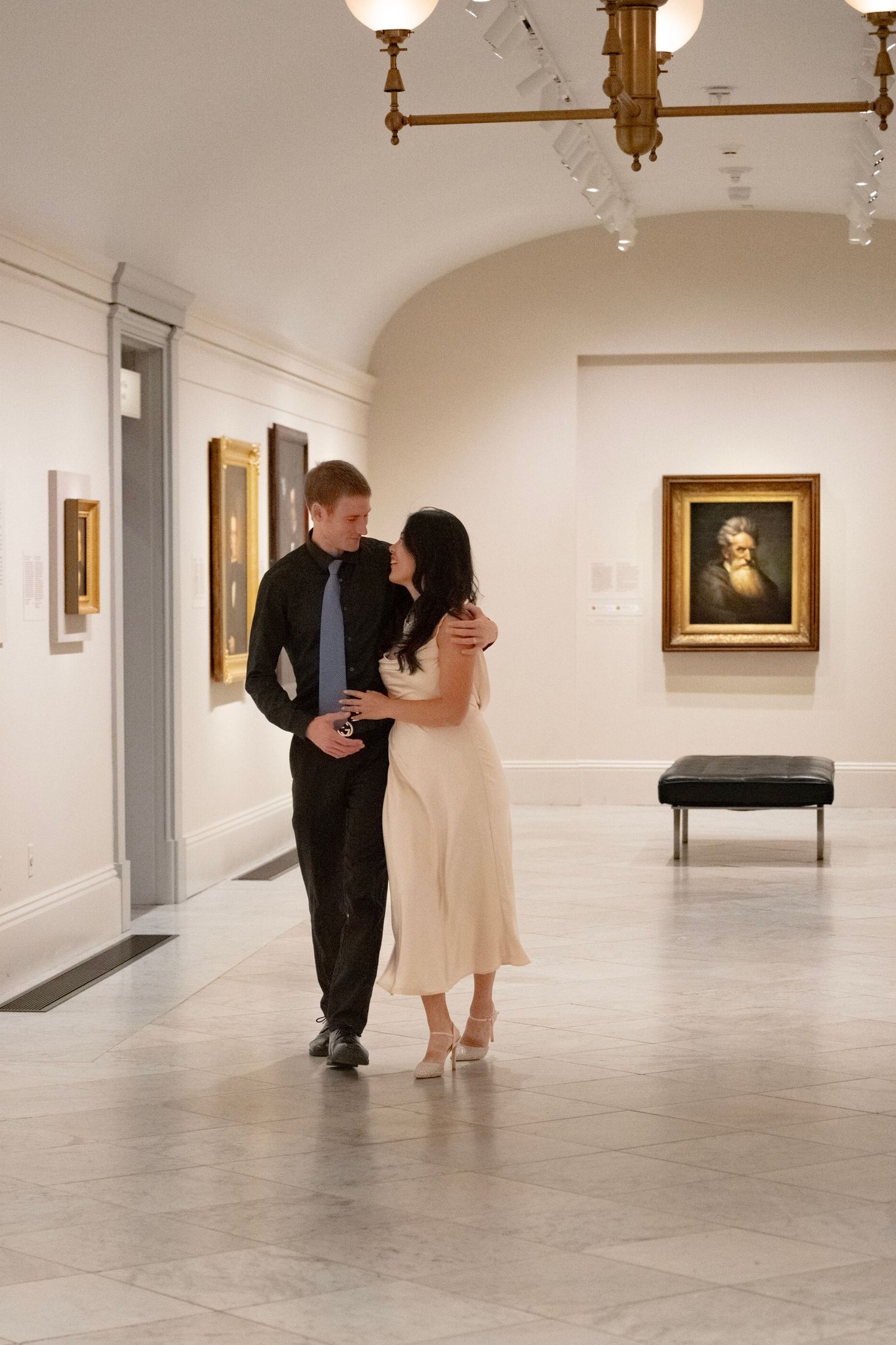 A couple walks through an elegant art gallery with white walls and framed paintings displayed in a spacious room.