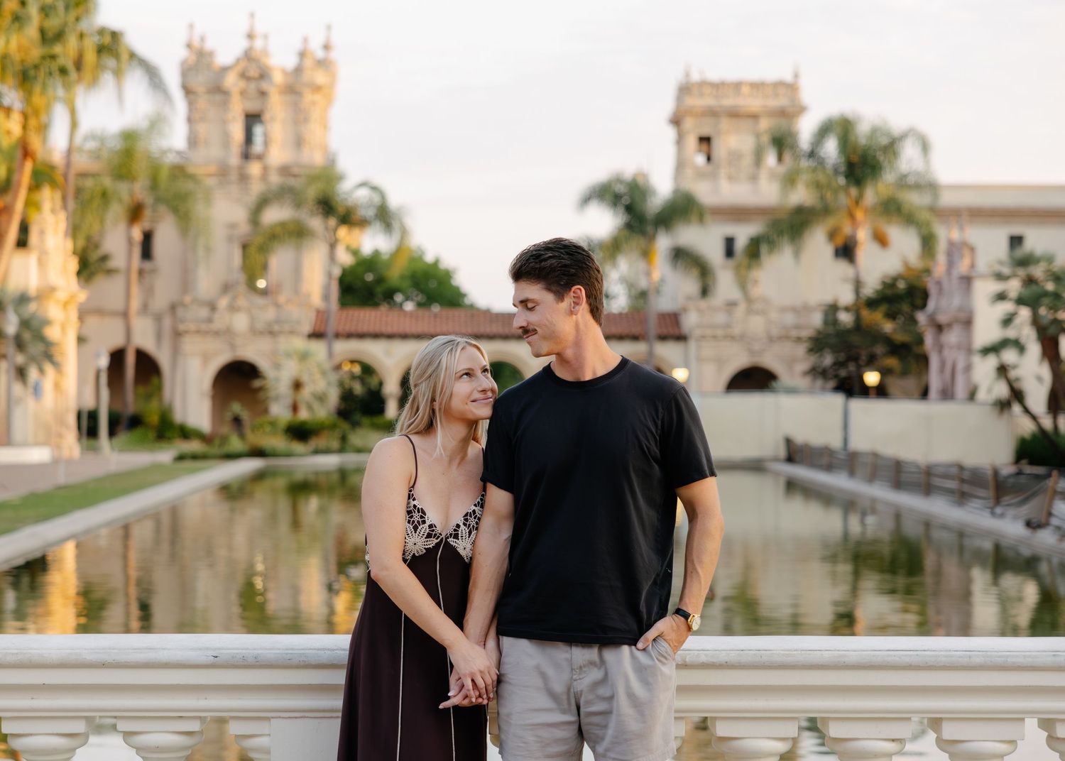 A couple poses together near a Spanish colonial building with palm trees at sunset.