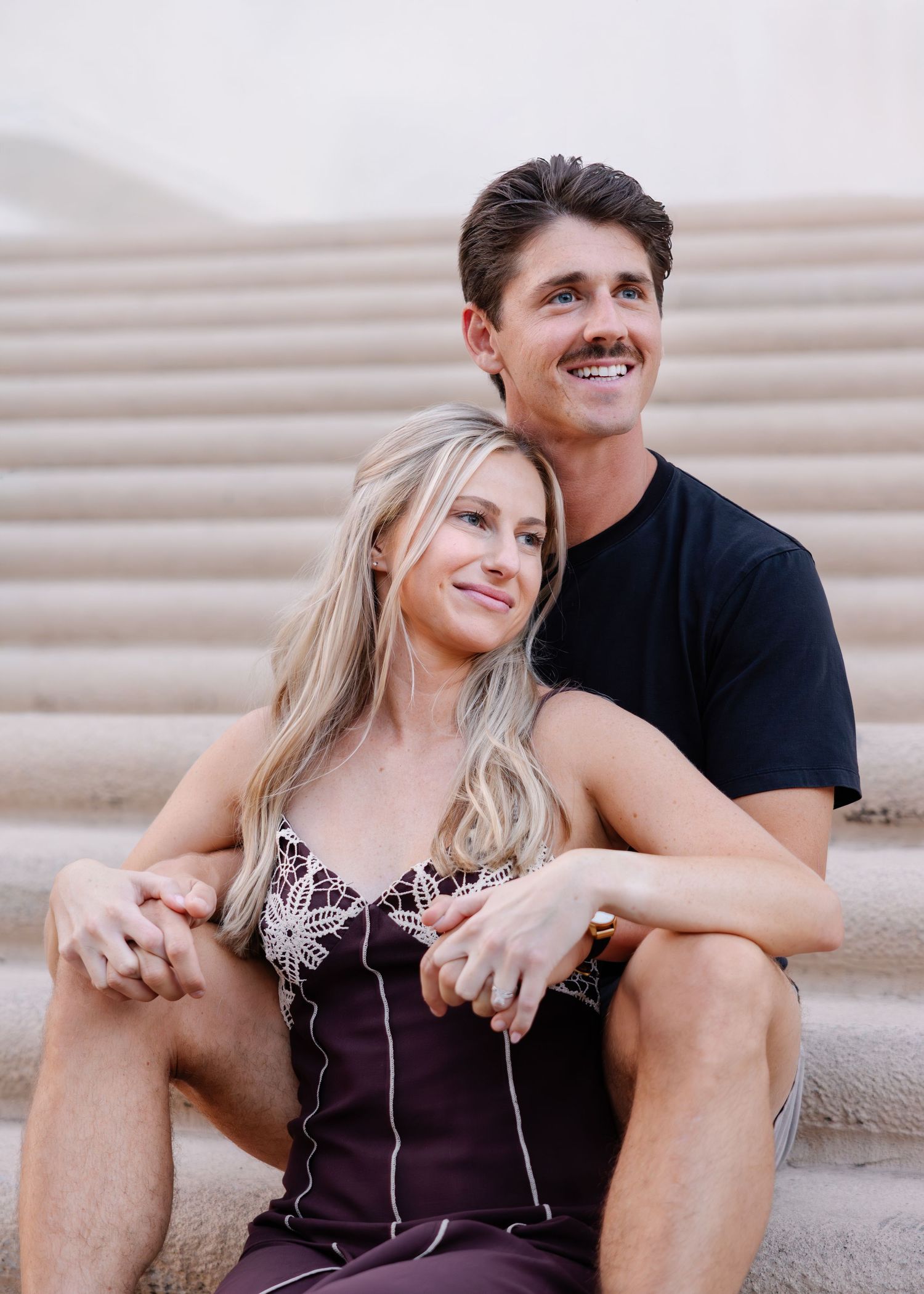 A romantic couple sits and laughs together on concrete steps on a sunny day.