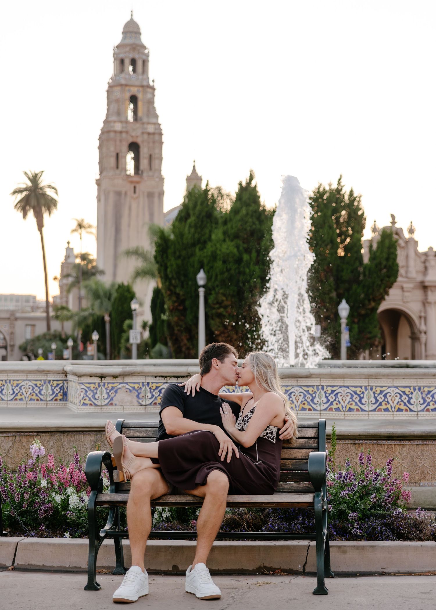 People relaxing on benches near a fountain with Spanish colonial architecture and palm trees in the background.