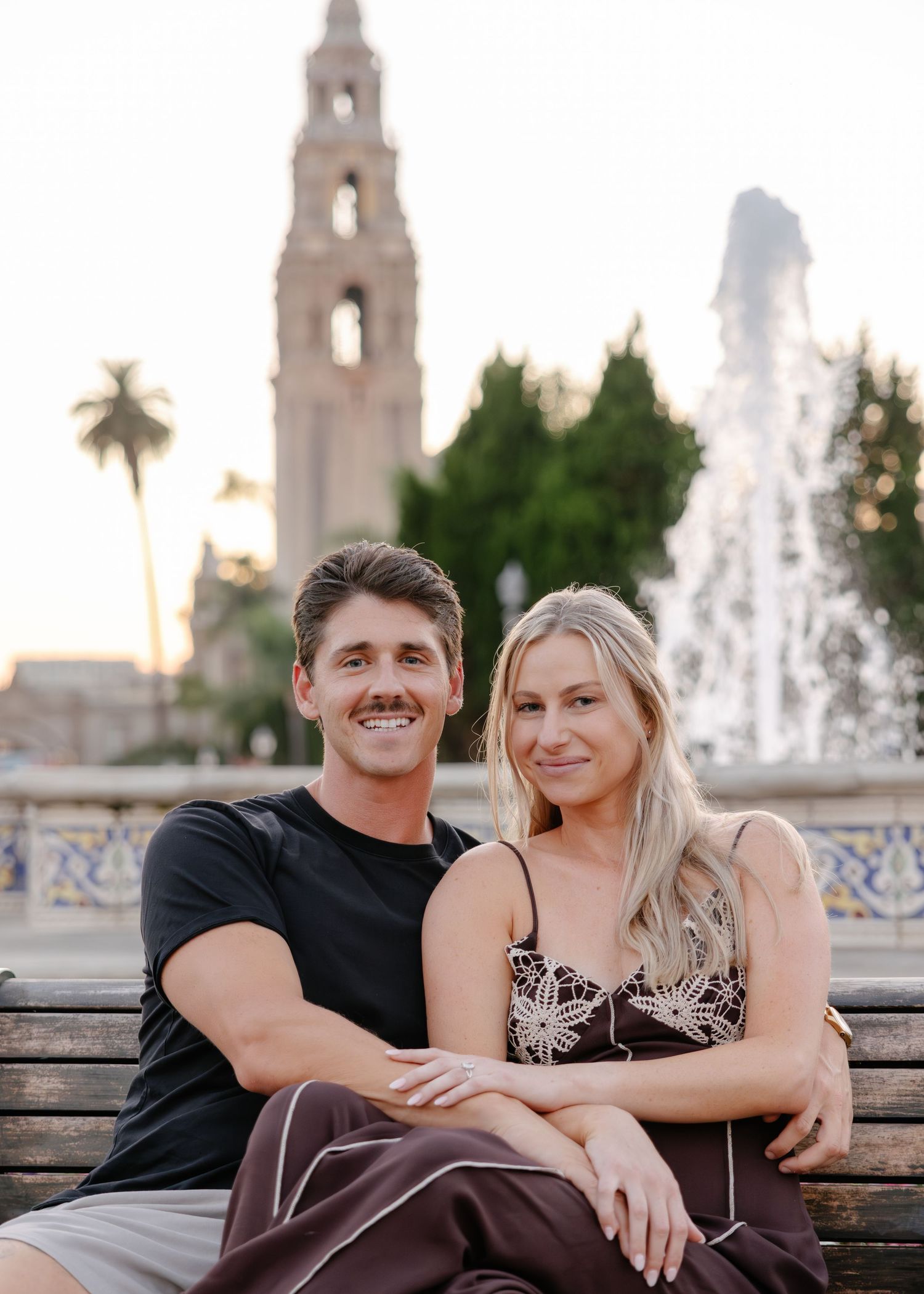 A couple sits together on a bench near a decorative fountain and Spanish colonial style bell tower at sunset.