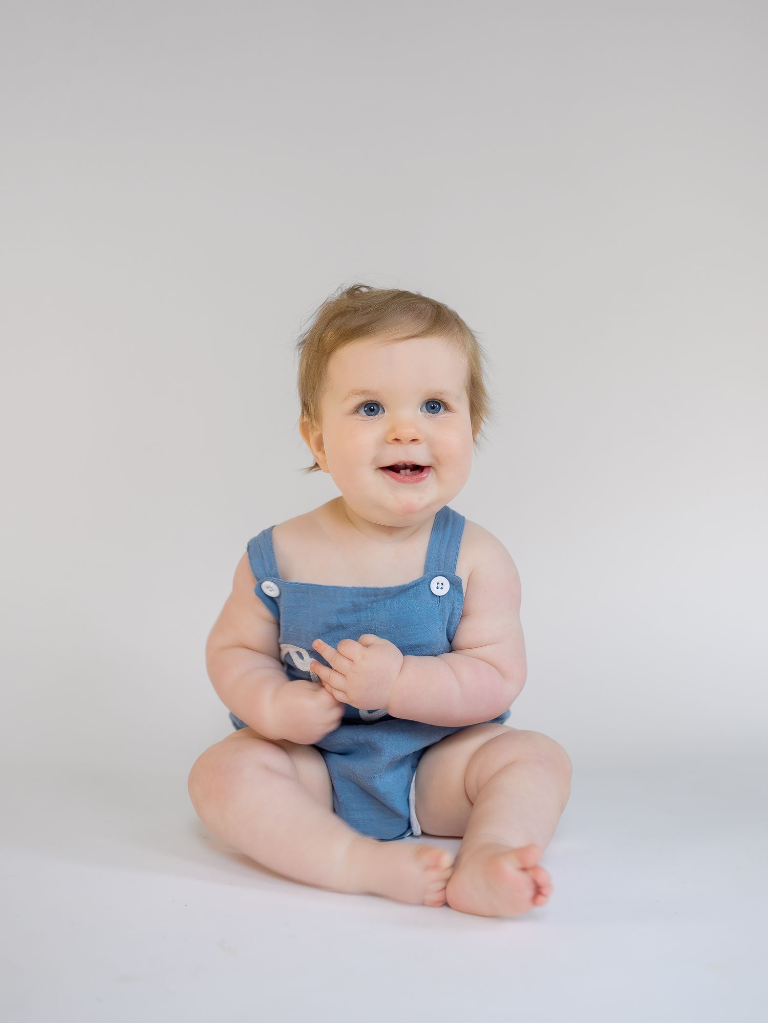 Smiling baby in blue romper sitting on white background, perfect for infant clothing photography.