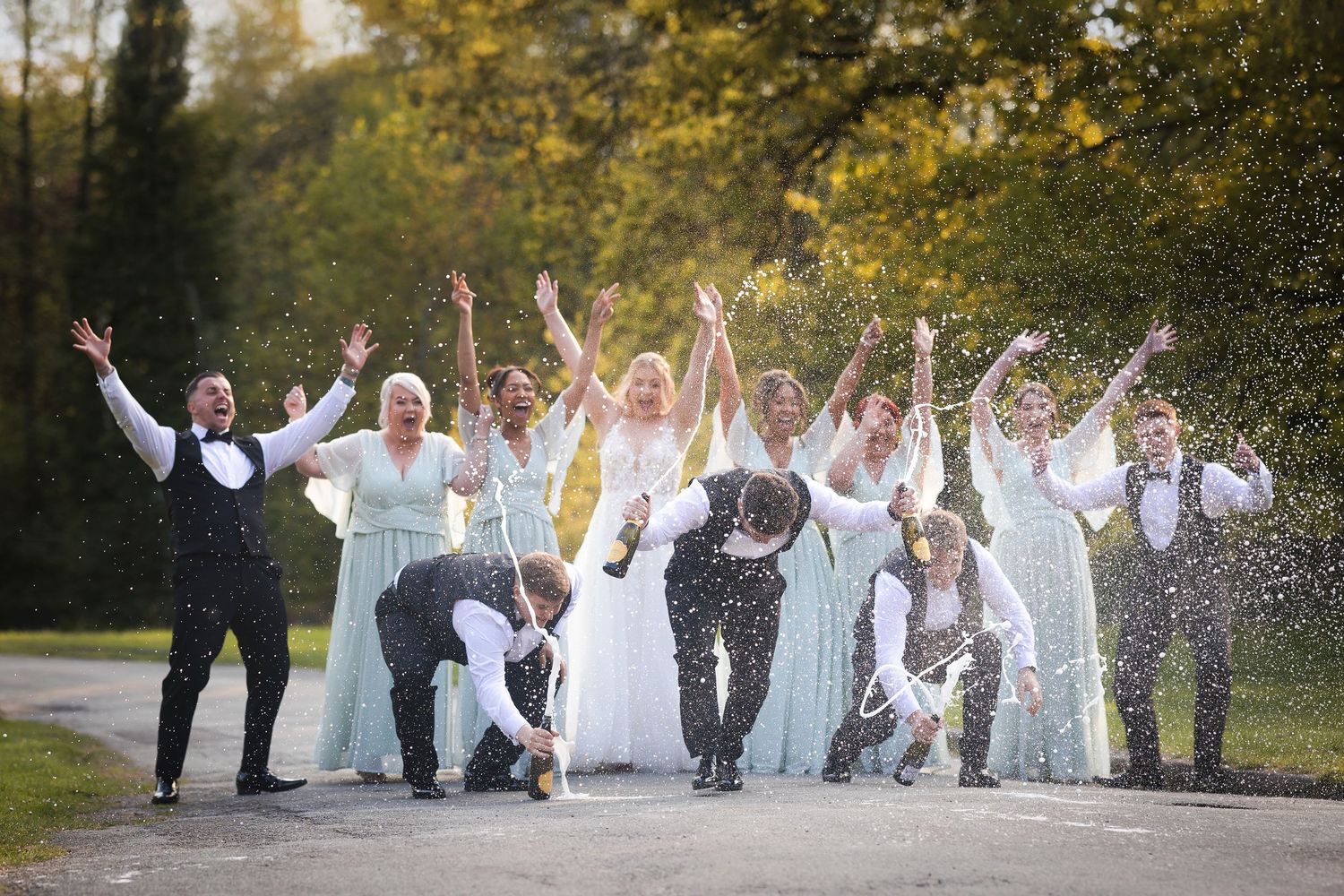 A group of celebrating people in white outfits jump joyfully in a garden setting during a special occasion.