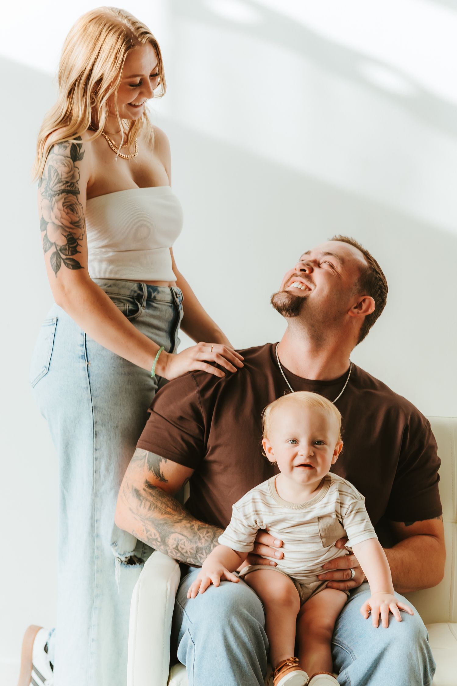 A couple poses against a beige wall, with a woman in a flowing blue floral dress and a man in a black polo shirt.