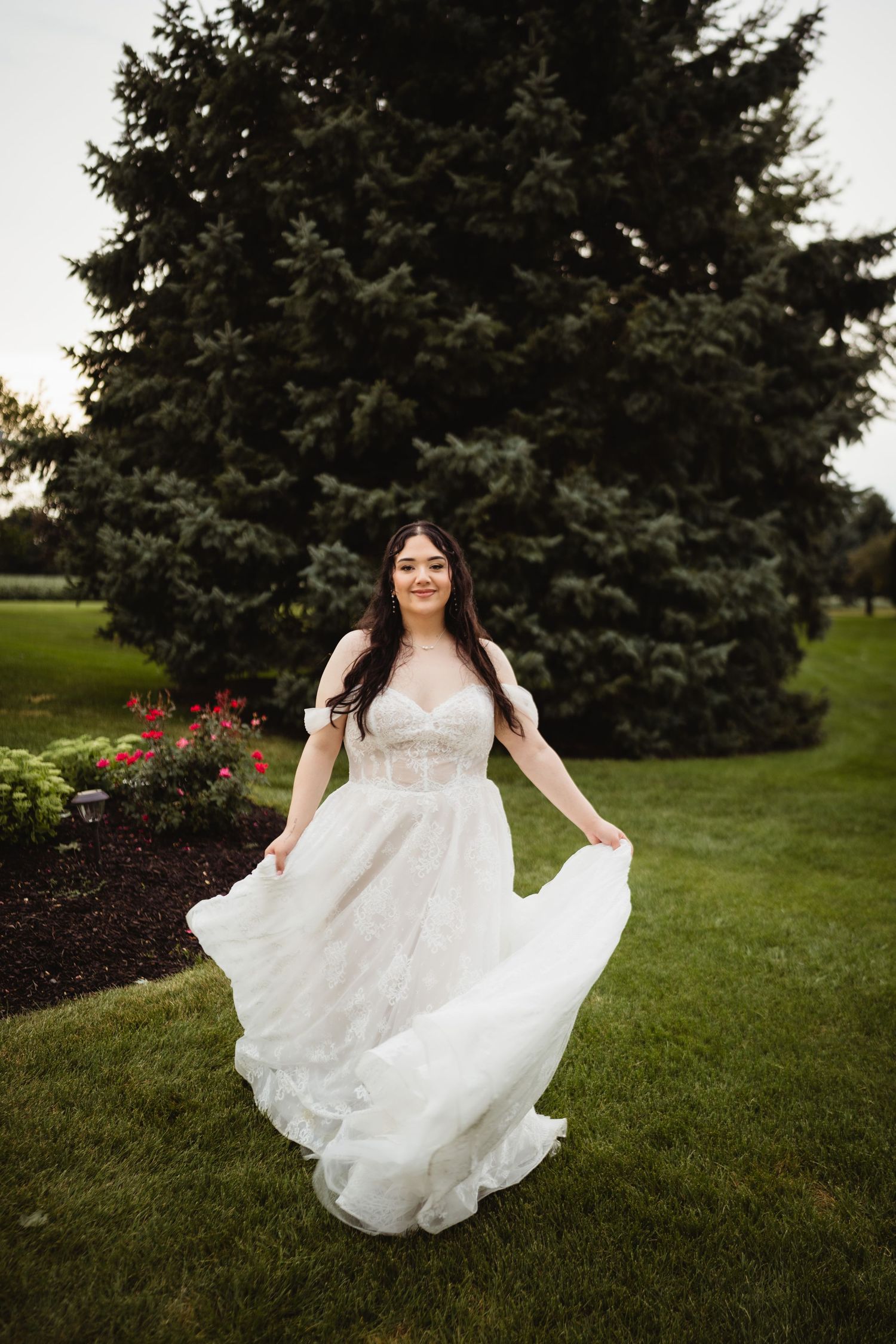 A person in a flowing white wedding dress twirls gracefully on a grassy lawn with evergreen trees in the background.