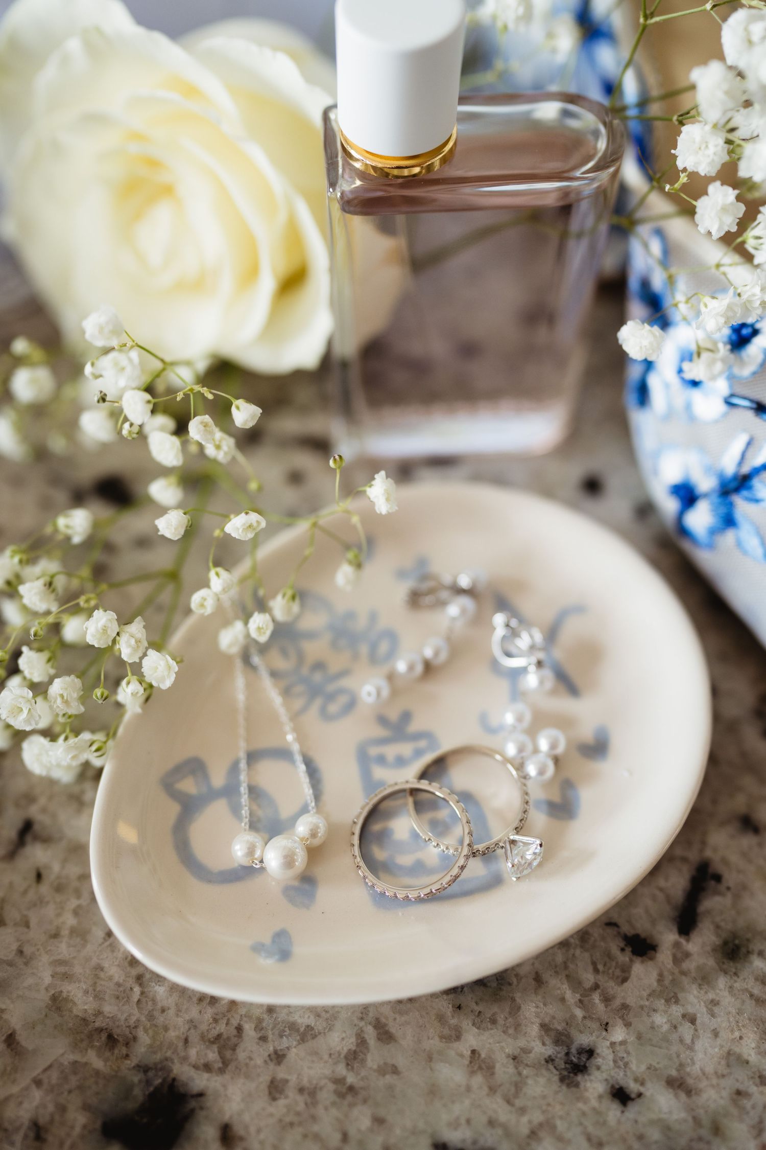 Wedding detail photo with white rose, delicate babys breath flowers, perfume bottle and jewelry on a decorative plate.
