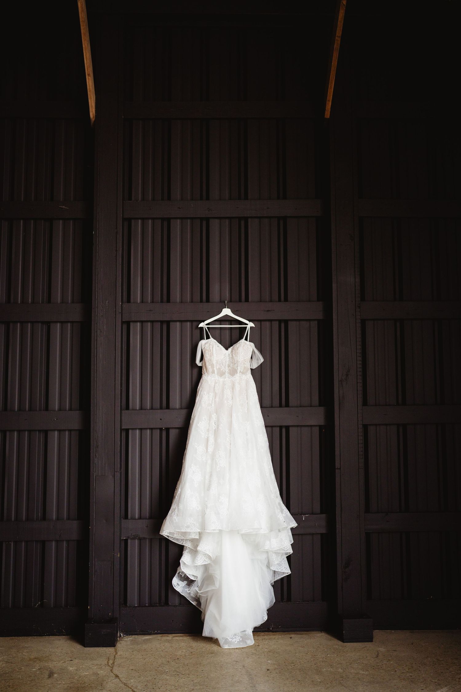 A white lace wedding dress hangs elegantly against a dark wooden wall, creating a striking contrast.