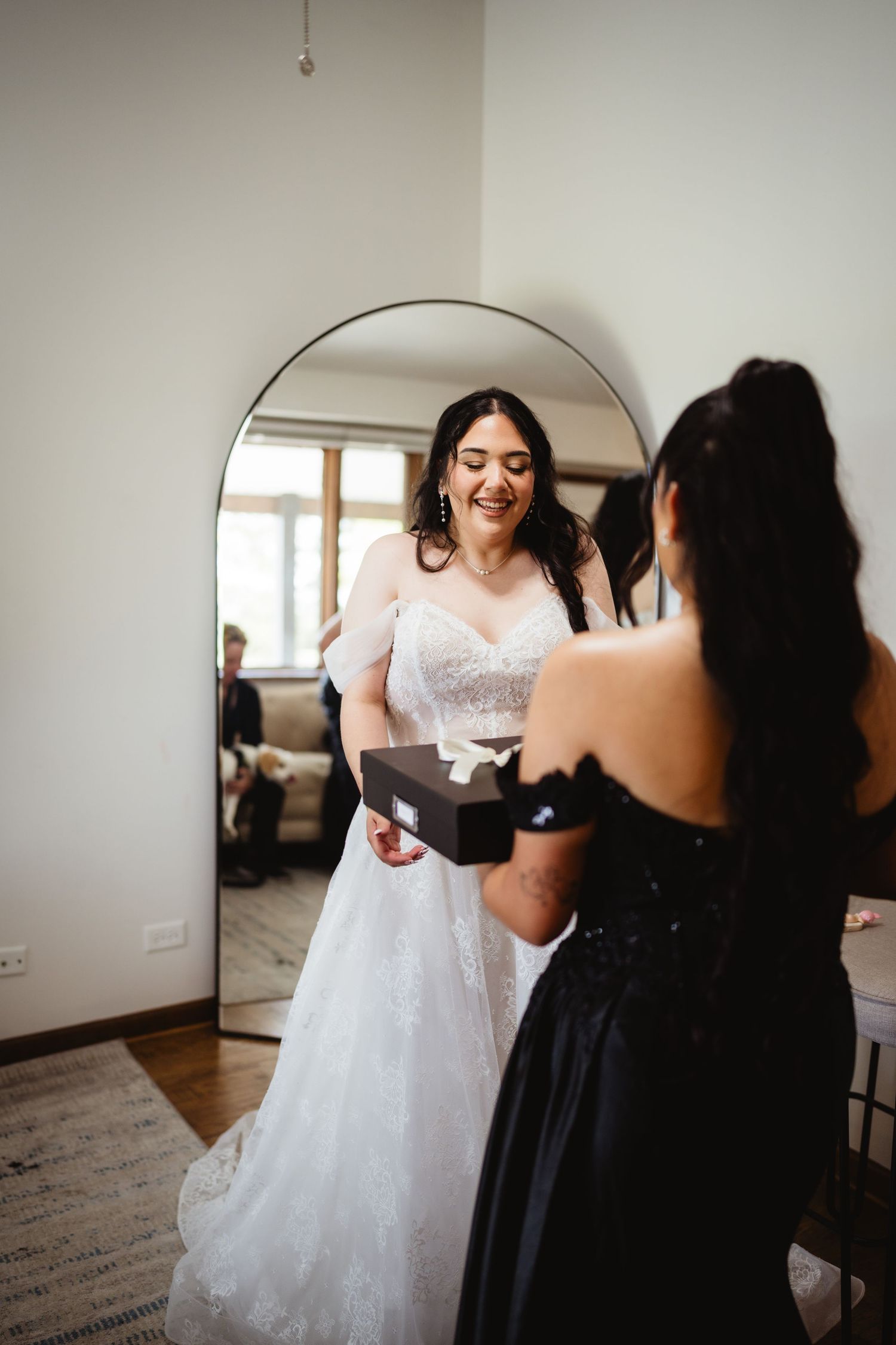 Joyful bride in white lace wedding dress looks at her reflection in arched mirror while wearing gown.