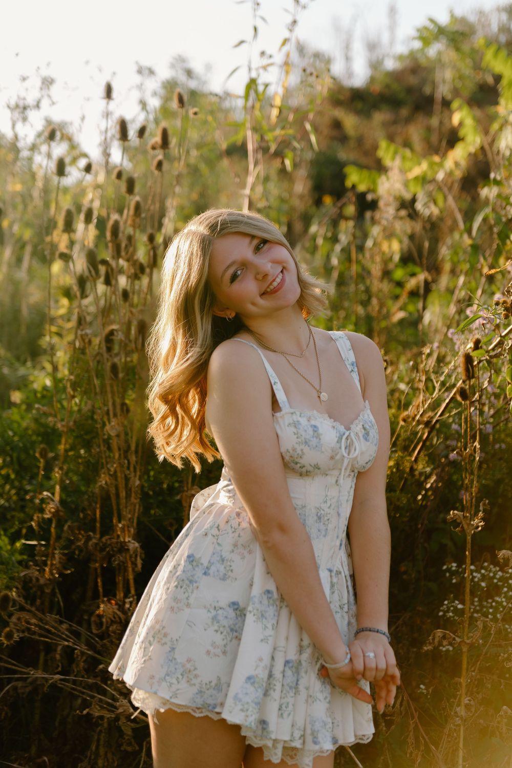 Smiling young woman in floral sundress standing in golden sunlit field with tall grass and wildflowers.