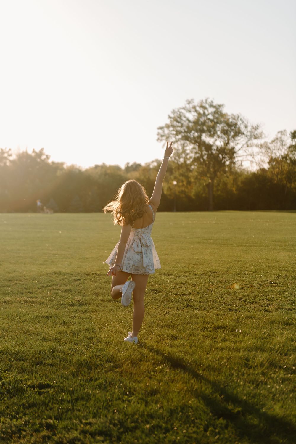 A carefree girl in a floral dress dances joyfully in a sunlit green park field with trees in the golden hour background.
