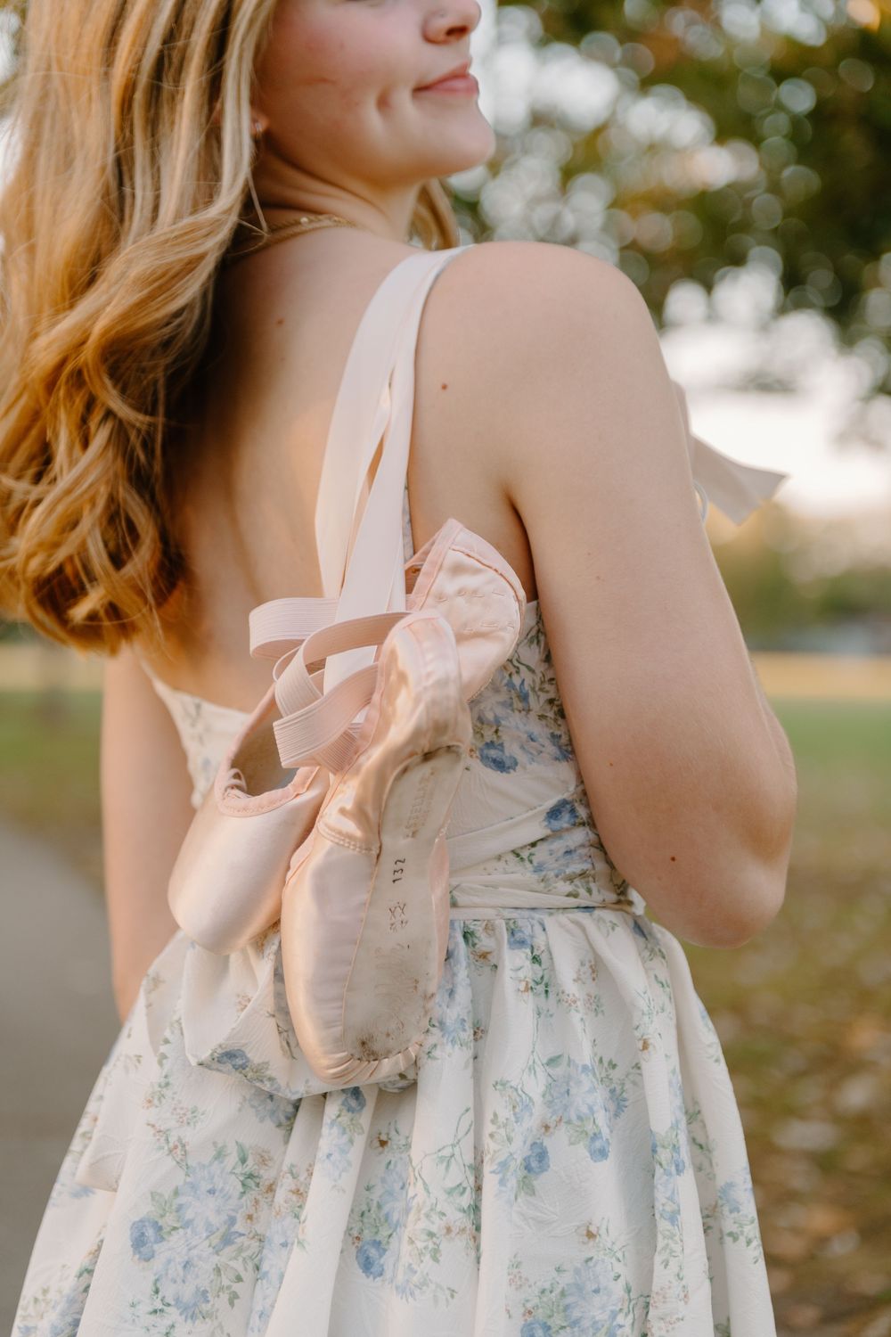 Blonde woman in a floral backless dress holding pink ballet pointe shoes outdoors in soft golden light.