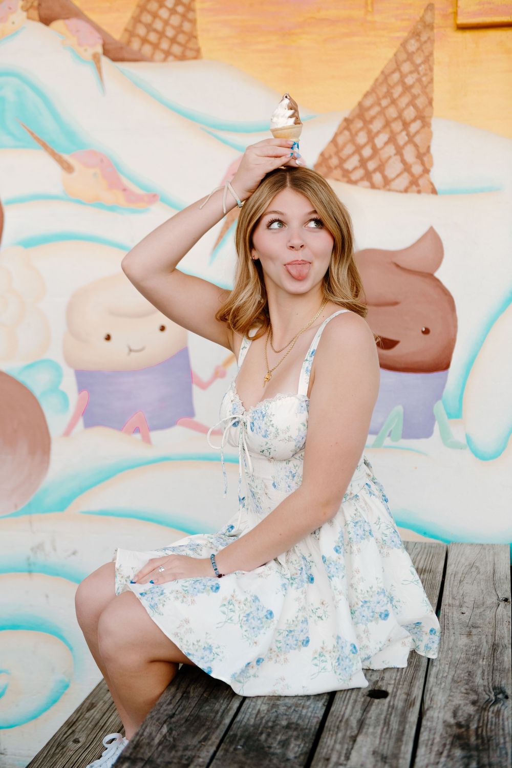 Young woman in floral dress holding ice cream cone on her head, sticking tongue out in front of colorful mural.