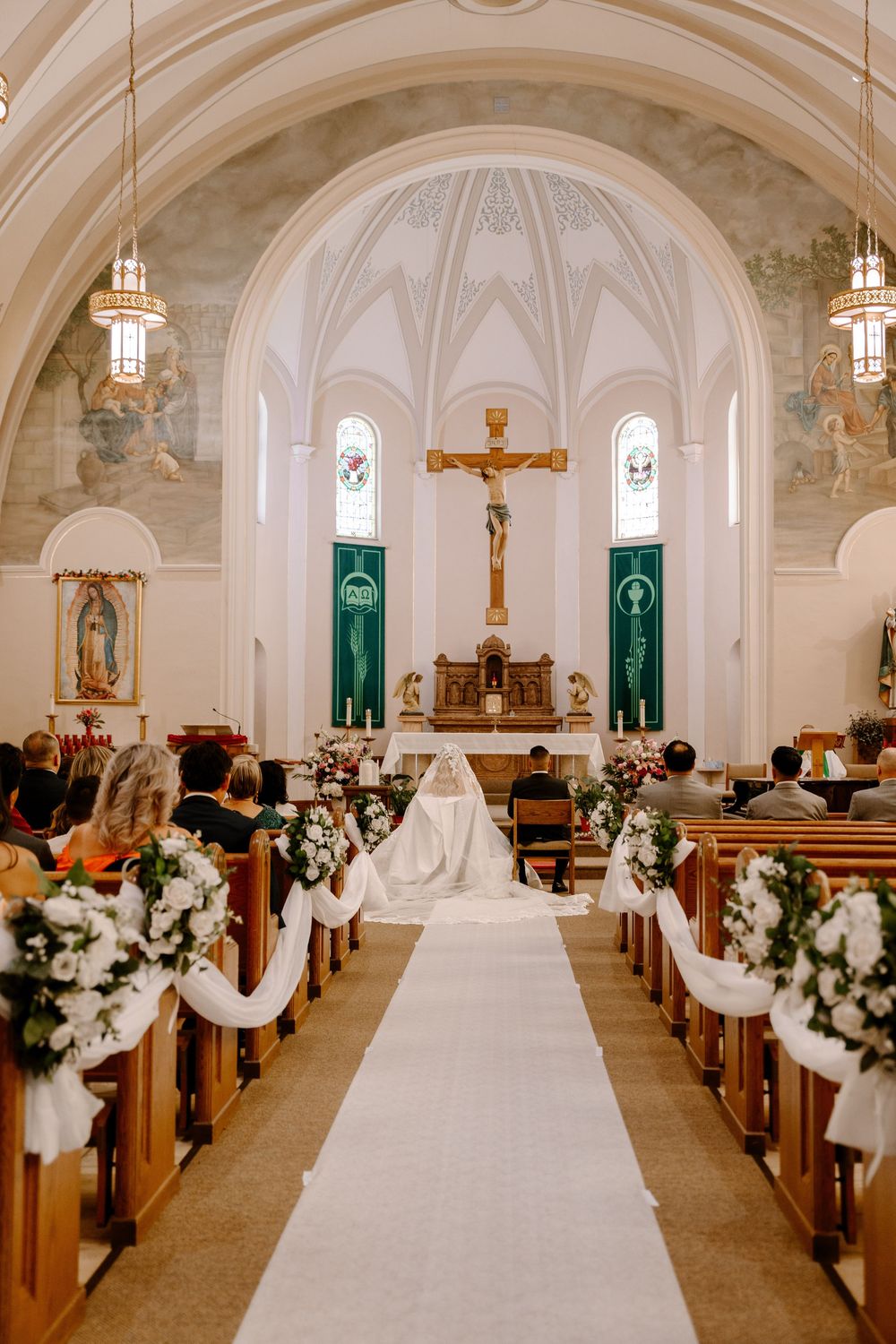 Interior of a traditional Catholic church during a wedding ceremony with white aisle runner and floral pew decorations.