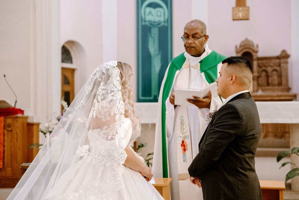 A bride and groom stand at the altar with a priest in green robes officiating their wedding ceremony in a church.