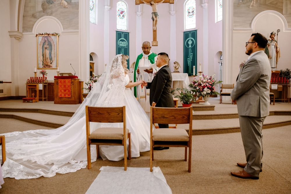 A wedding ceremony takes place inside a traditional church with a white-robed priest officiating at the altar.