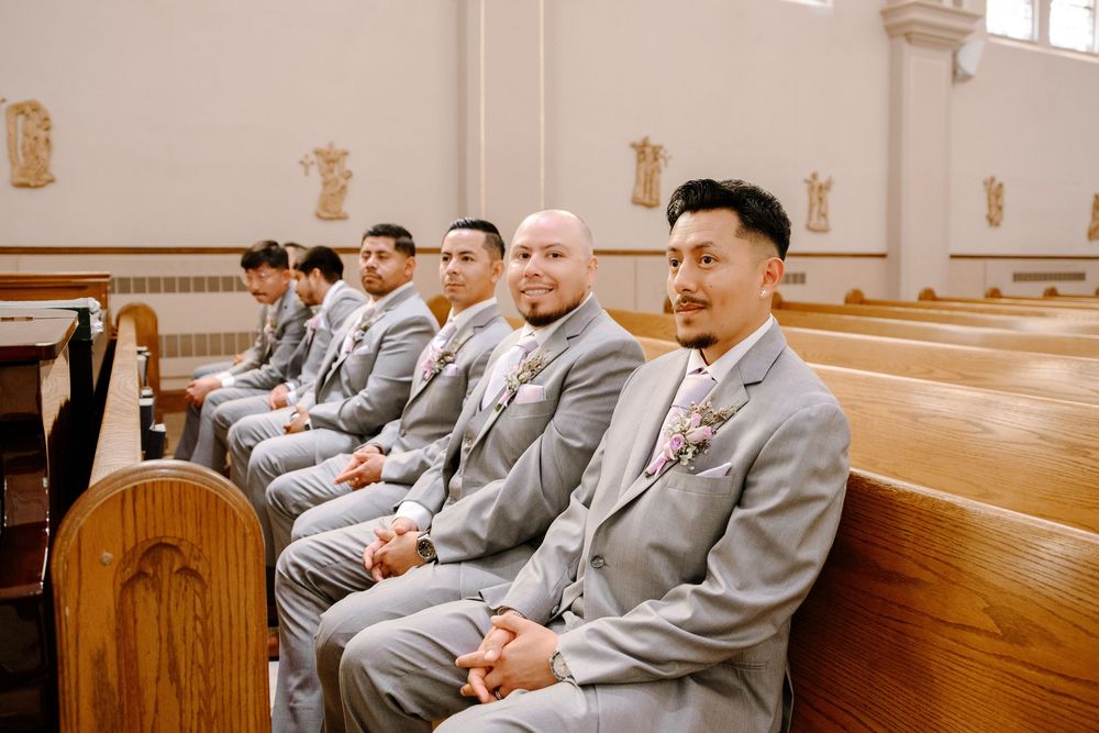 Groomsmen in matching gray suits sit in wooden church pews during a wedding ceremony.