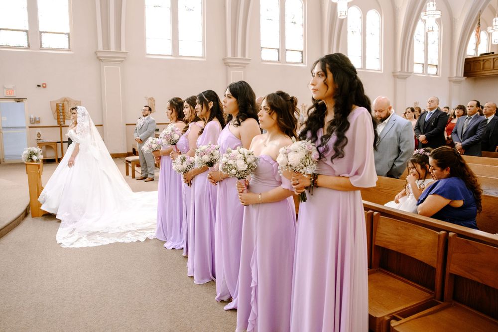 Bridesmaids in flowing lilac dresses stand in line watching bride at church wedding ceremony.