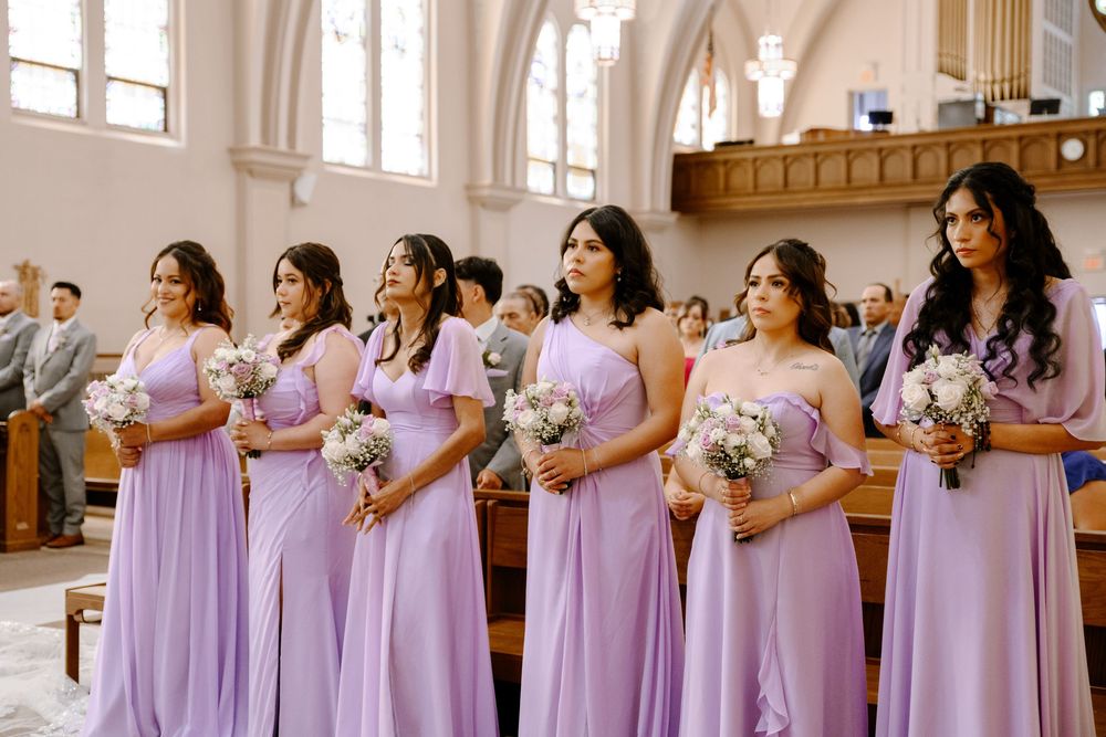 Bridesmaids in flowing lilac purple dresses line up holding white floral bouquets inside a sunlit church.