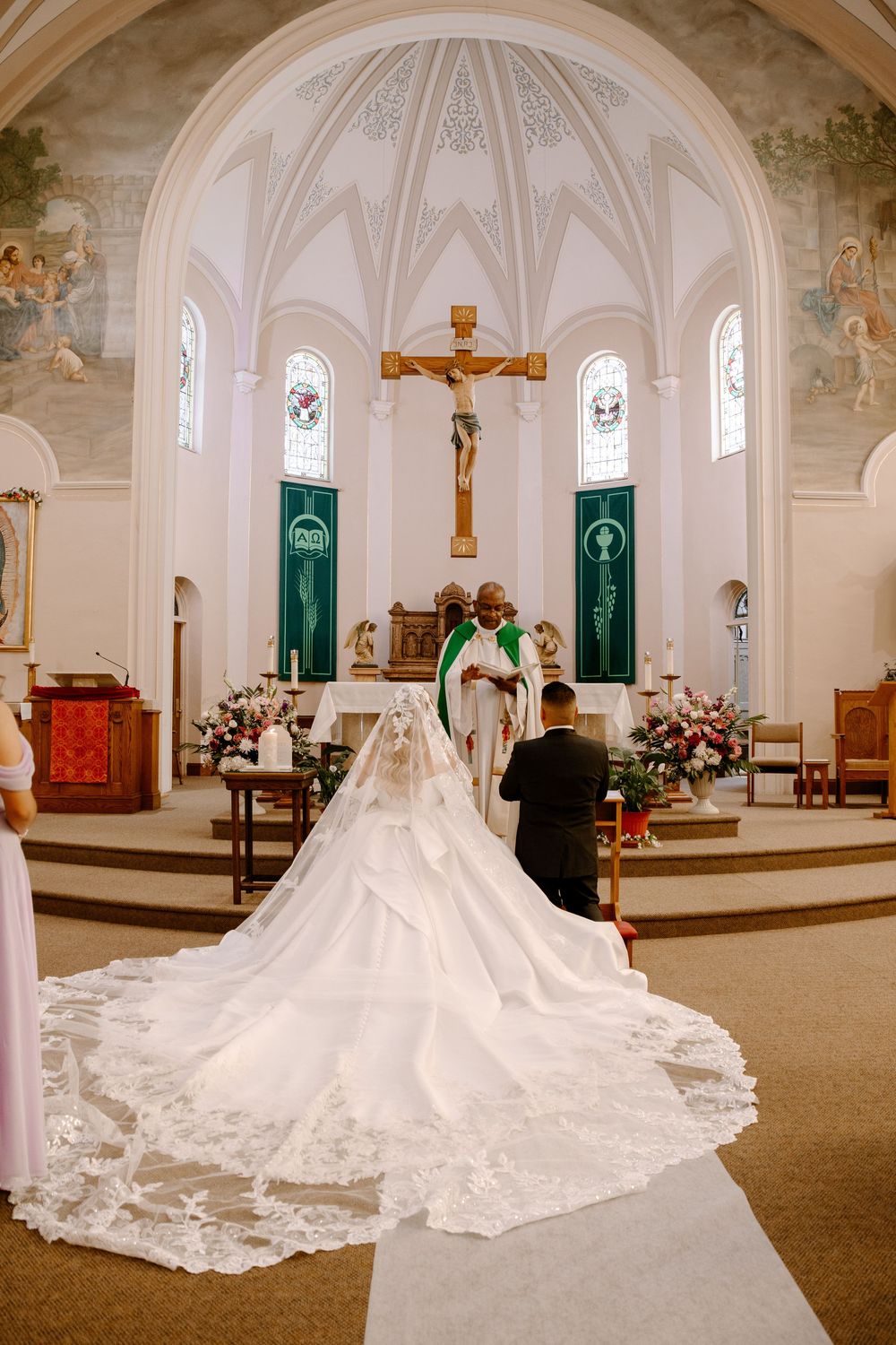 A wedding ceremony takes place in a beautiful Catholic church with vaulted ceilings and a long flowing lace bridal train.