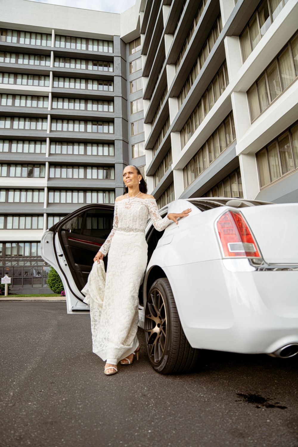 A white wedding dress and luxury white sedan car in front of a modern apartment building.