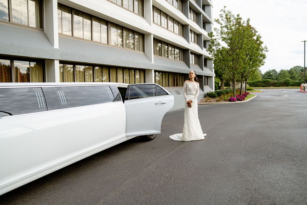 A white stretch limousine parked outside a modern building with someone in a flowing white dress standing nearby.