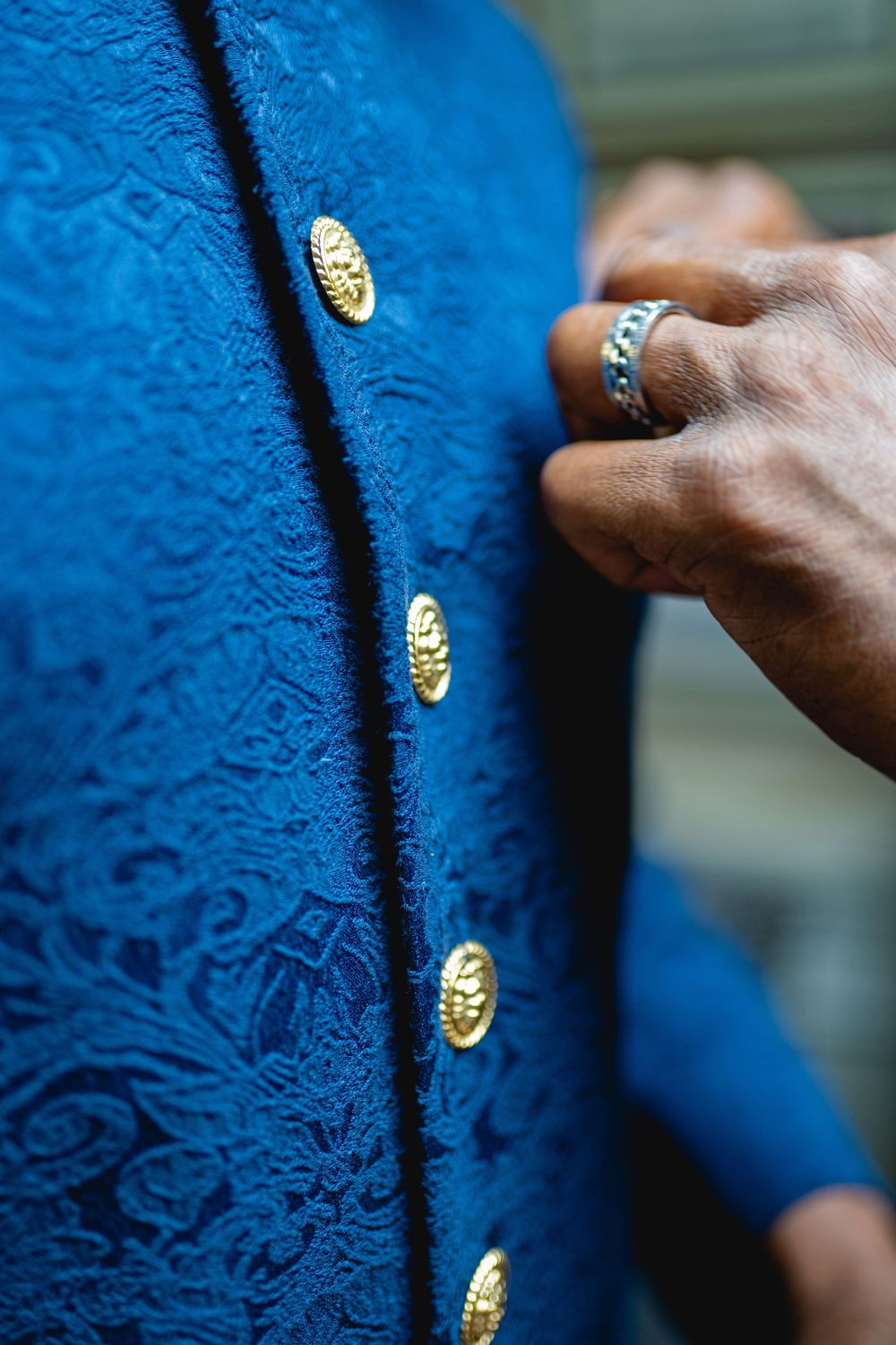 Close-up of a hand wearing turquoise ring adjusting gold buttons on an ornate blue brocade jacket. Wedding at Westbury Manor