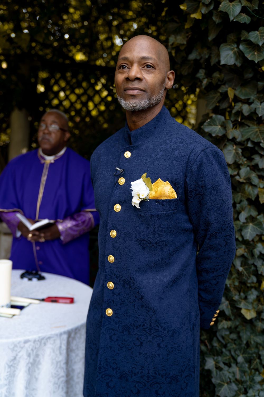 A figure in a navy blue formal jacket with gold buttons stands in front of greenery at an outdoor event. Wedding at Westbury Manor