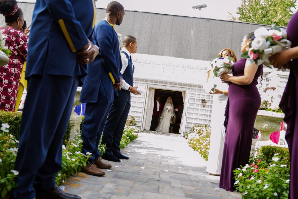 Guests in formal attire line the entrance path of a white stone building during an outdoor wedding ceremony. Wedding at Westbury Manor