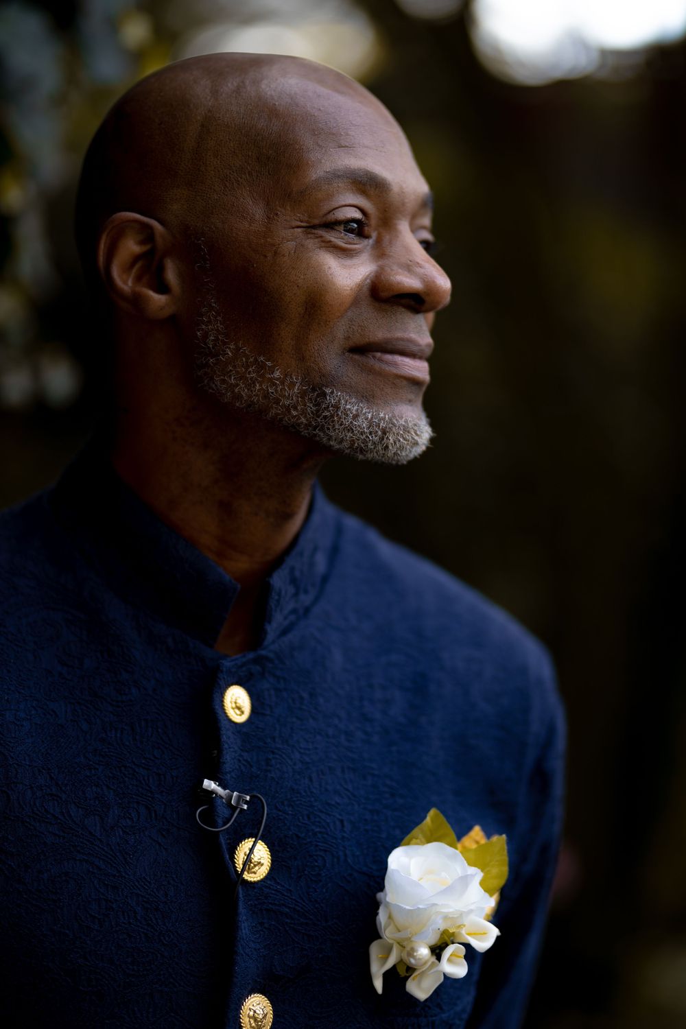 A well-dressed person in a navy jacket with gold buttons and white boutonniere smiles while looking to the side. Wedding at Westbury Manor