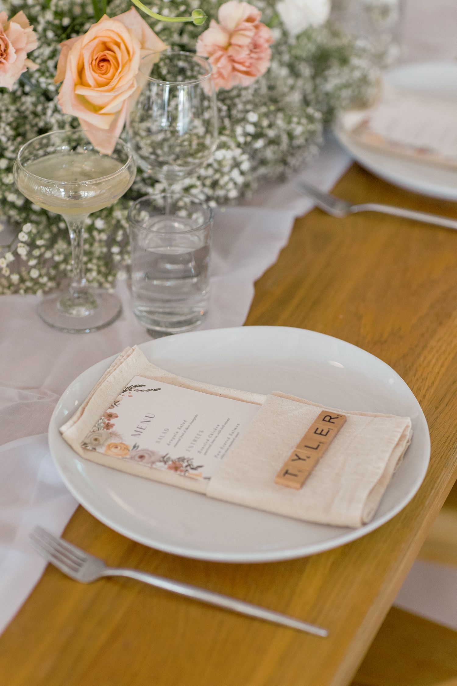 Place setting at a wedding reception with peach roses, baby's breath flowers, and vintage menu card on wooden table.