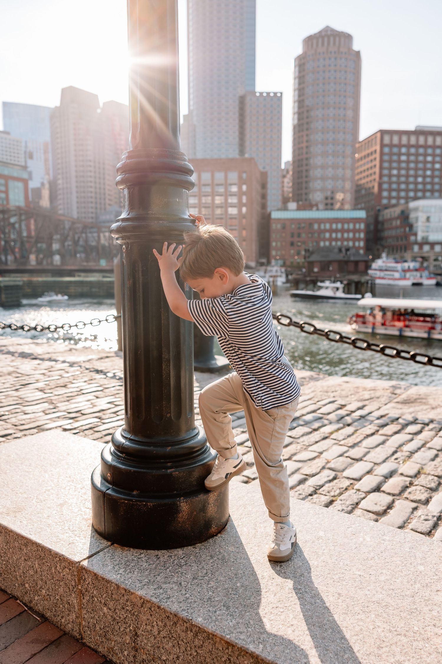 Child in striped shirt plays by historic harbor bollard on cobblestone waterfront with Boston skyline in background.