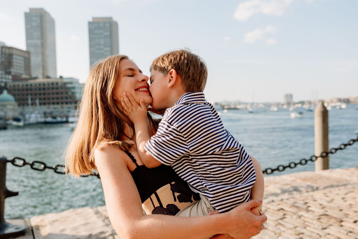 A romantic couple shares tender moments by the waterfront with Boston Harbor and boats in the background.