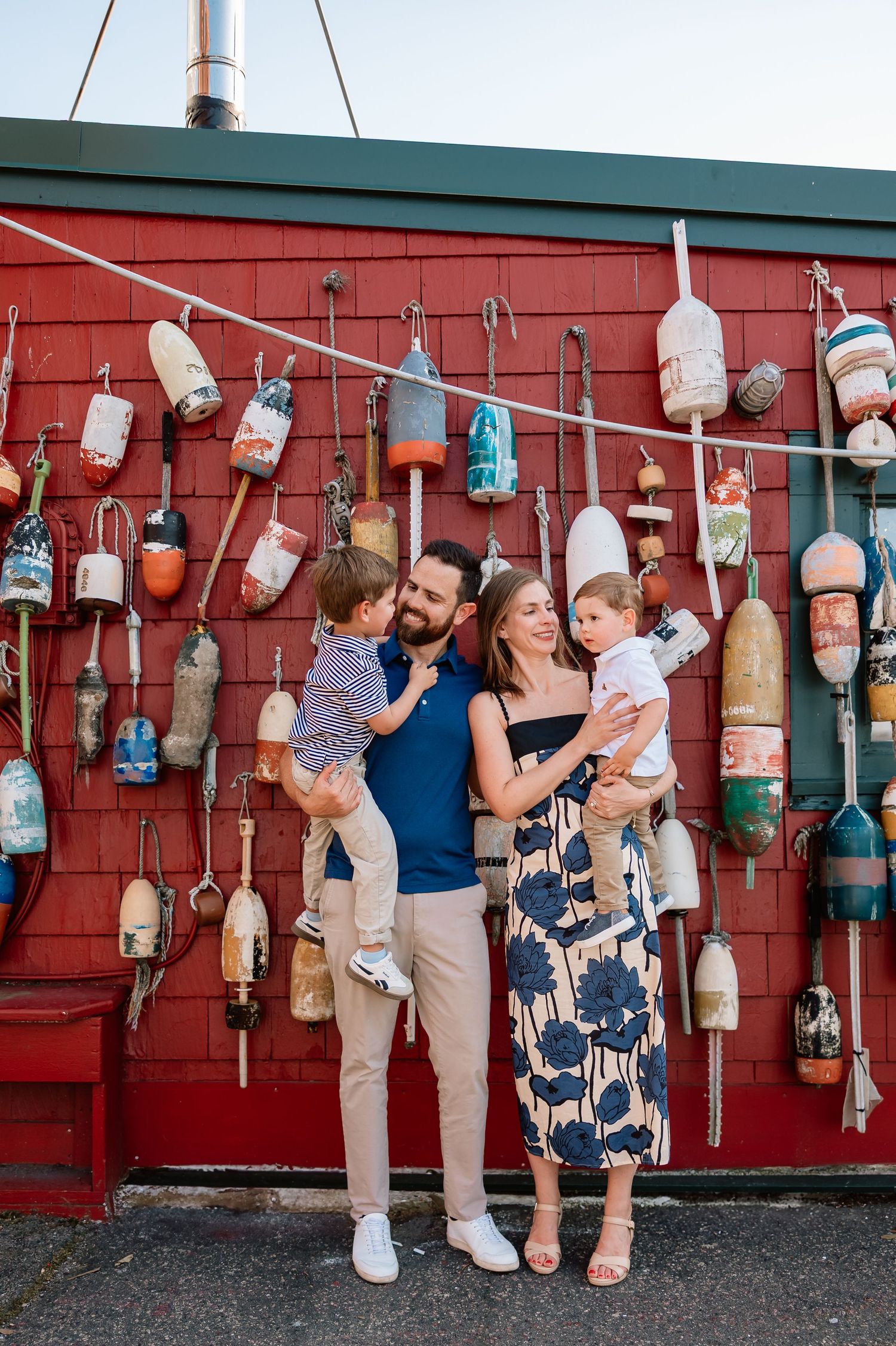 Family in coordinated outfits poses against red barn wall decorated with hanging buoys and nautical decor.