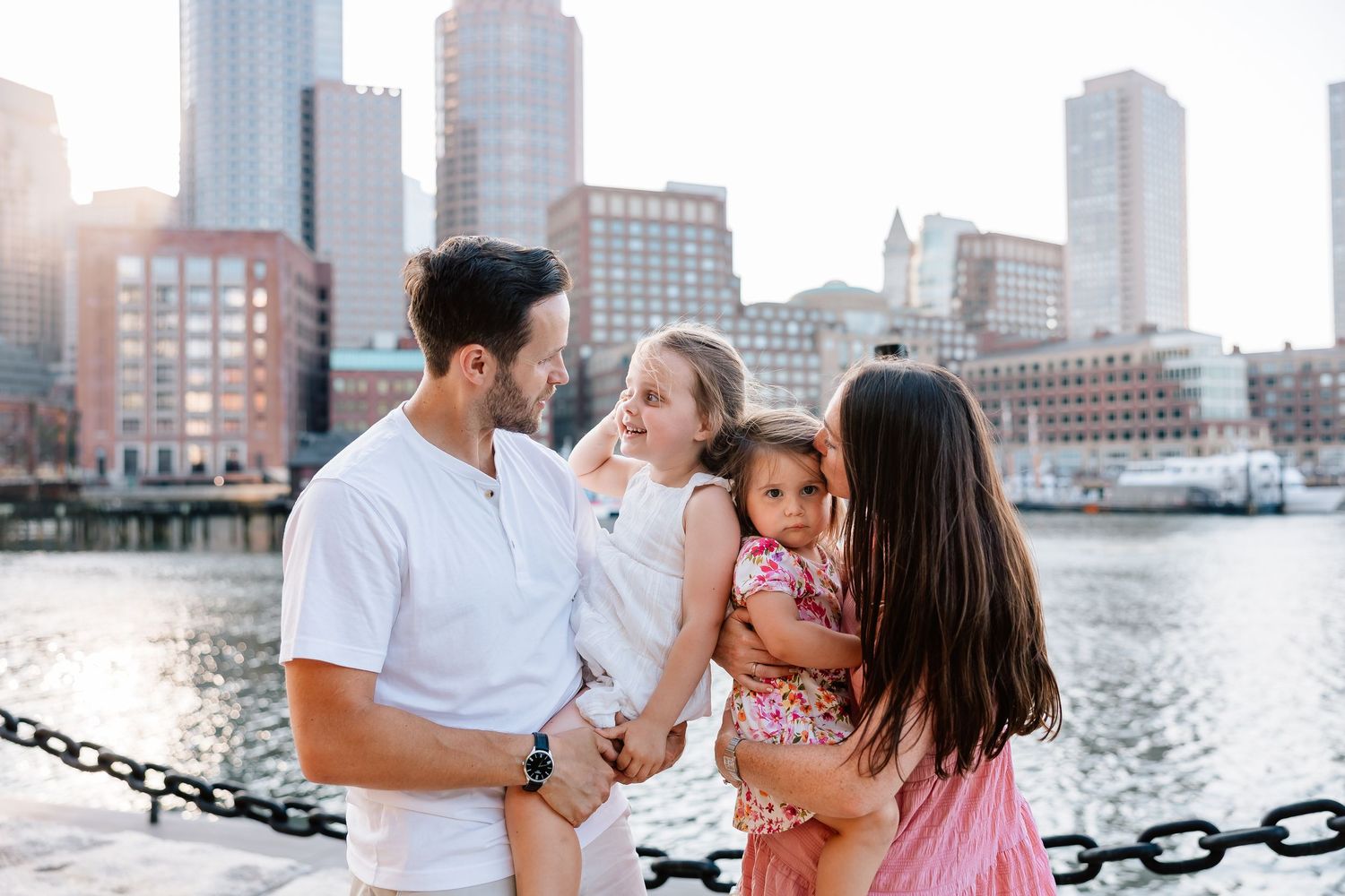 Family enjoying a waterfront stroll along a city harbor with urban buildings in the background during sunset.