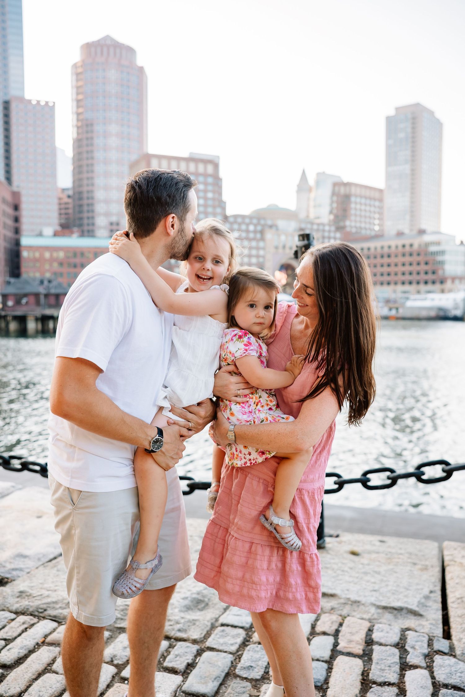 Family sharing tender moments together on cobblestone waterfront with city skyline in background during sunset.