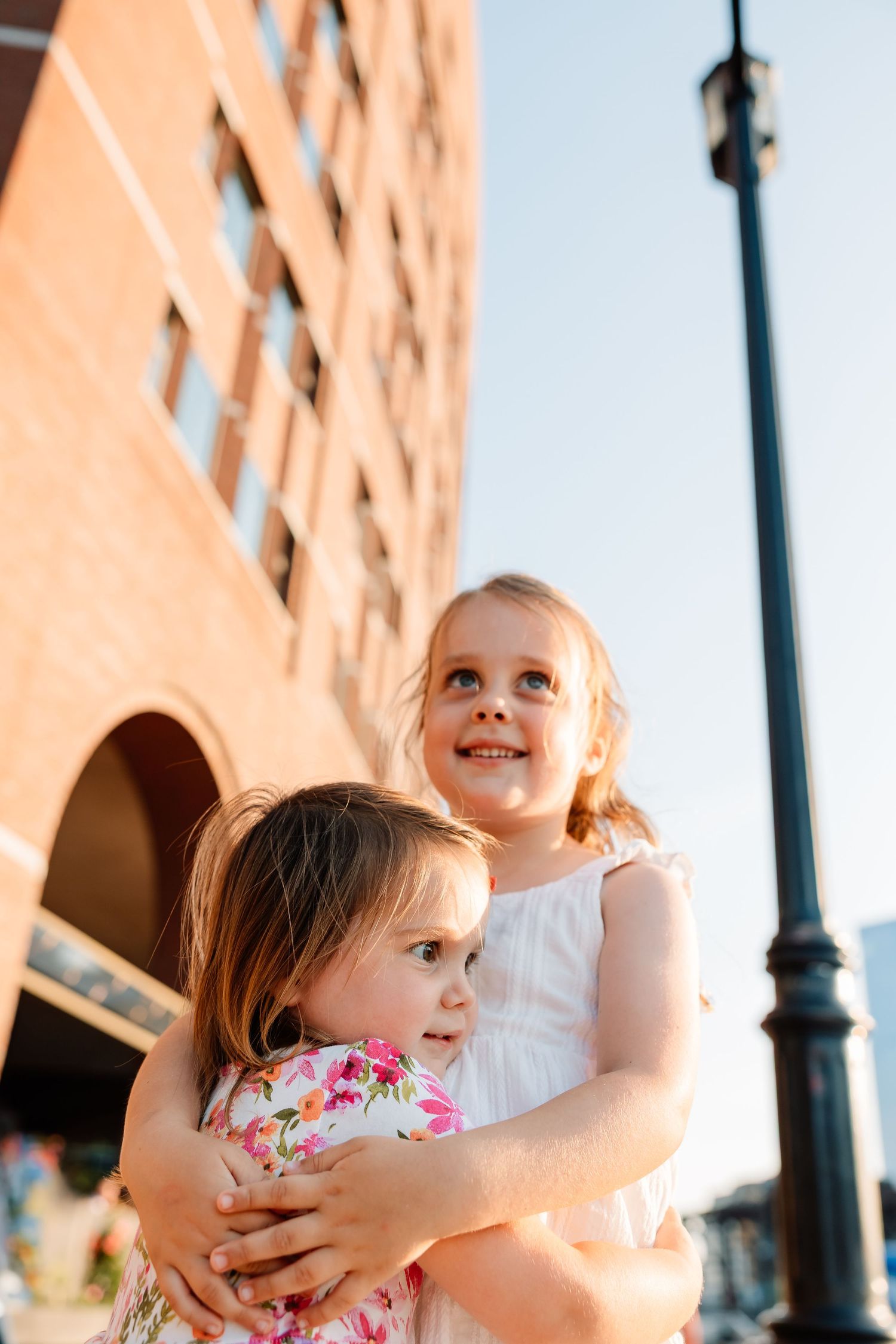 Two young children embrace and smile while standing near a building with an ornate lamp post at sunset.