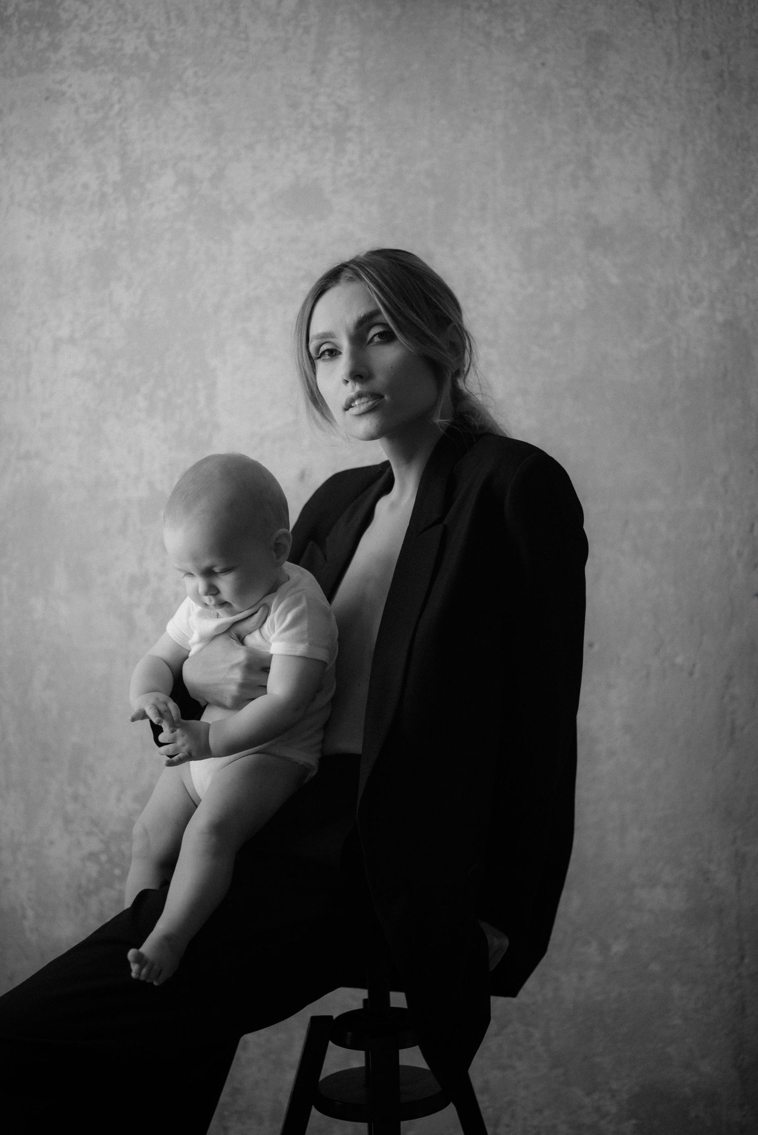 Black and white studio portrait of a woman in dark blazer seated on stool holding baby in white outfit.