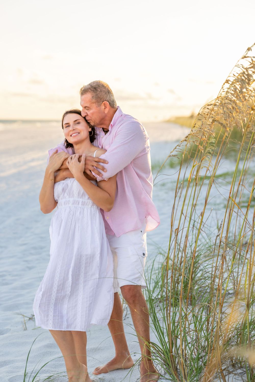 A couple shares an intimate moment together on a sandy beach with sea oats swaying in the golden sunlight.