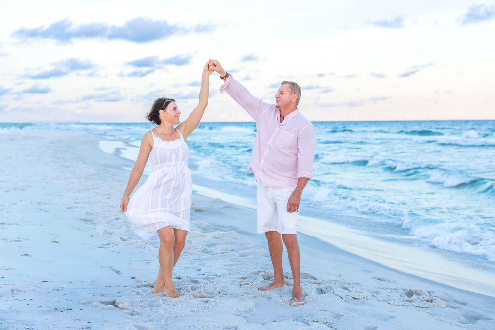 A couple dances playfully together on a white sandy beach at sunset with waves crashing in the background.