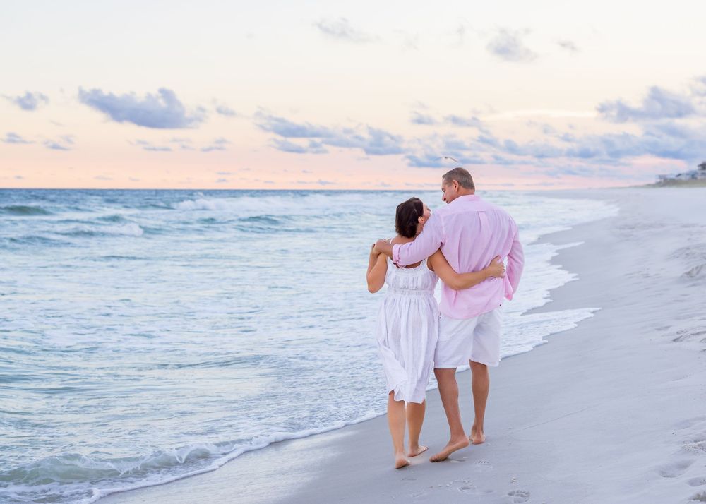 A couple in white clothing walks along a peaceful beach at sunset with waves rolling onto the shore.