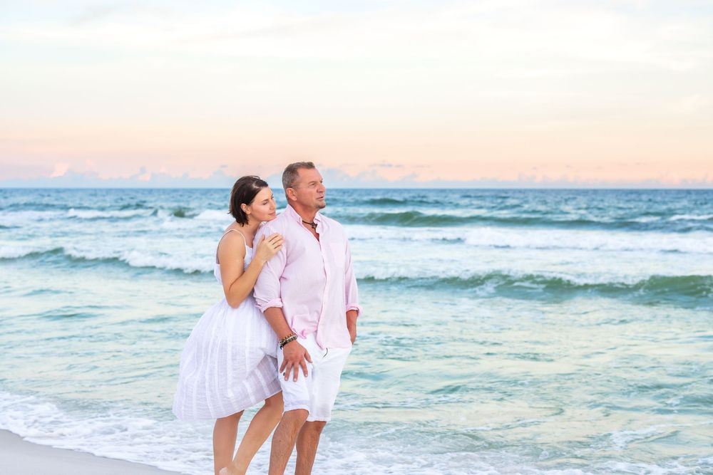 A couple in white attire embraces while walking along a beach at sunset with turquoise waves rolling in.