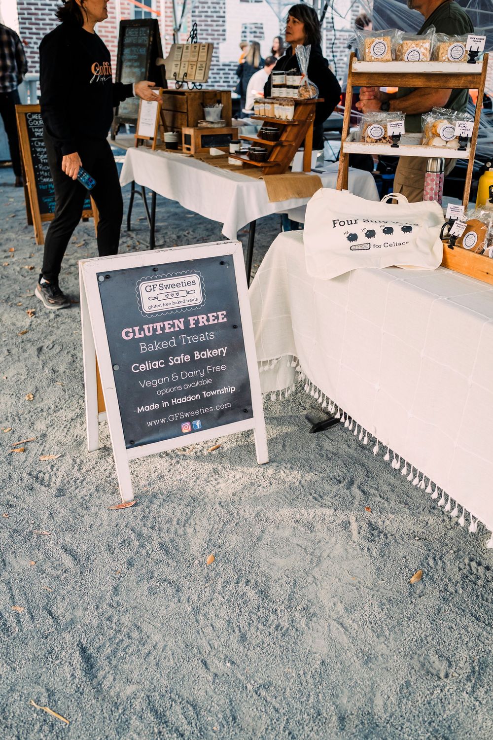 A-frame chalkboard sign advertising gluten free options at an artisanal market vendor stand with display shelves.